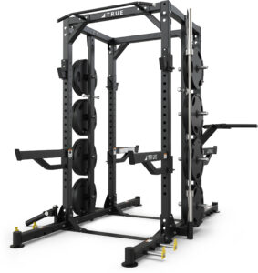 A black power rack with multiple weight plates, safety bars, pull-up bar, and attachments for strength training, set on a white background. The rack is labeled TRUE and has storage pegs for organizing weights.