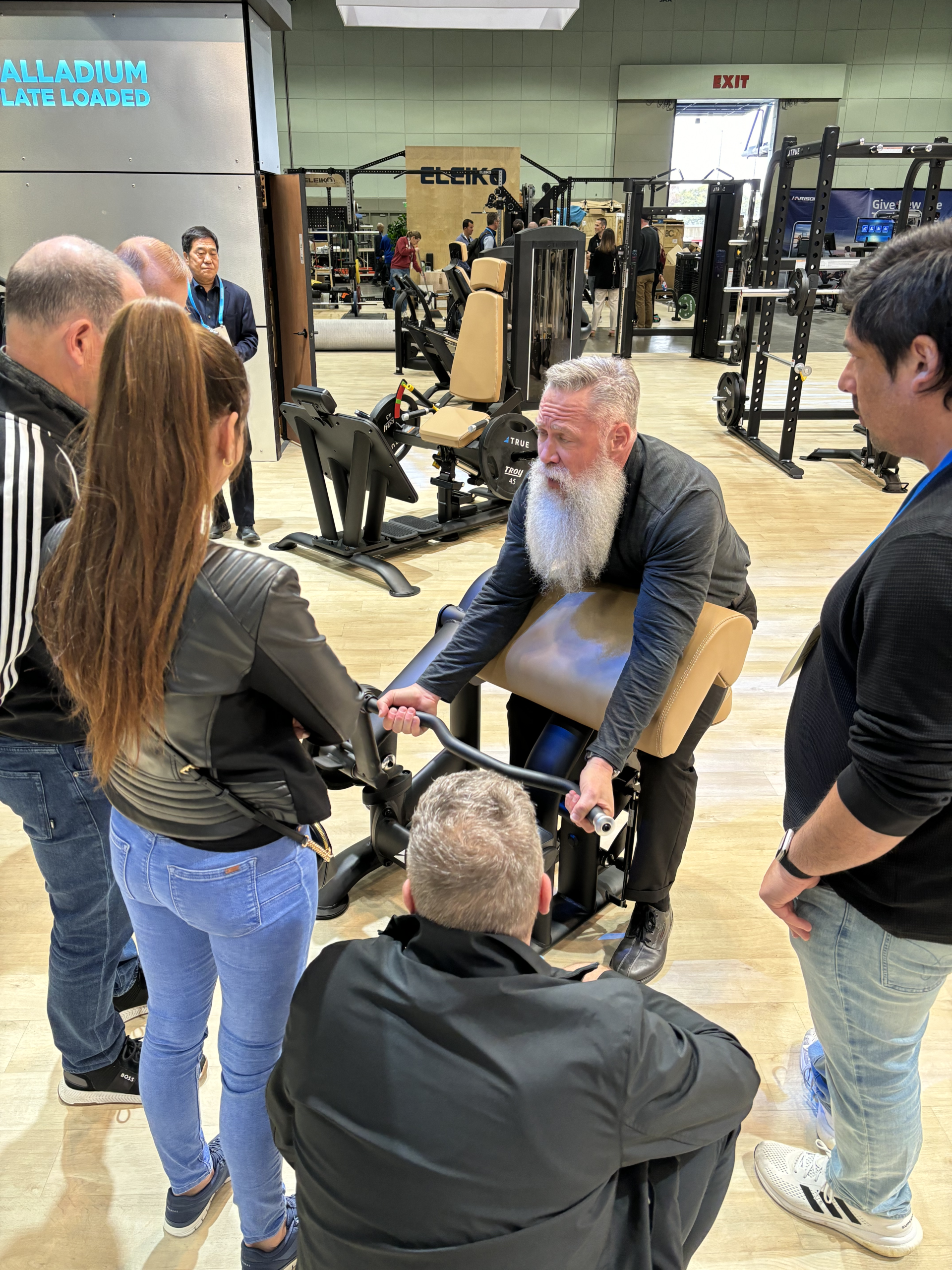 A bearded man demonstrates a fitness machine to a small group of people in a gym equipment showroom, with various exercise machines and weights visible in the background.