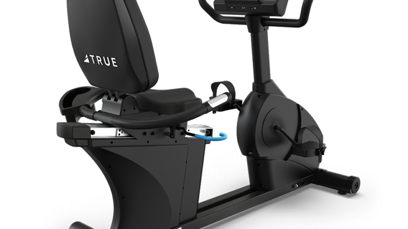 A black recumbent exercise bike with a digital touchscreen display, adjustable seat with backrest, and pedals. The brand name TRUE is visible on the bike’s frame and seat back.