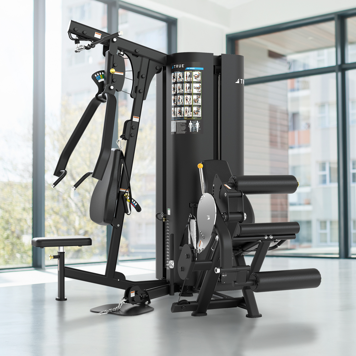 A multi-station gym machine with various attachments, including a leg extension, chest press, and pull-down handles, positioned in a bright room with large windows and a view of surrounding buildings.