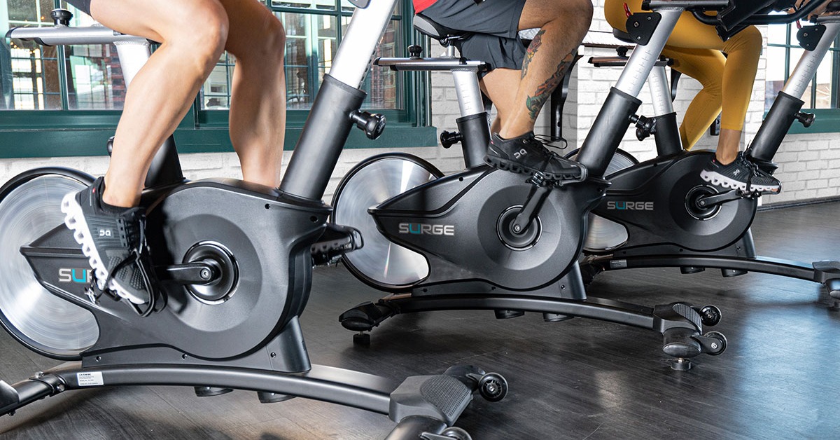 Three people pedal on black Surge stationary exercise bikes. They wear athletic clothing and lean over the handlebars.